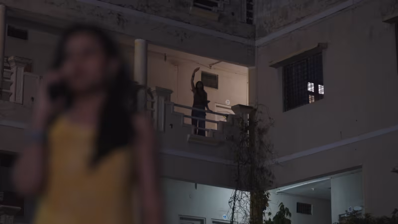 She waves from the balcony into the courtyard while a blurred figure watches from the foreground