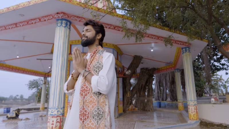 A still from Mere Dil Mein Bhole Baba: a man in white standing with folded hands at a riverside temple